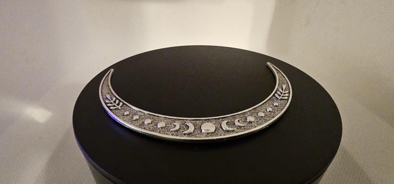 Puede incluir: Una luna creciente plateada con una superficie texturizada y un dise&ntilde;o de peque&ntilde;os c&iacute;rculos y lunas crecientes. La luna est&aacute; sobre una superficie negra.