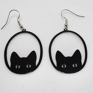 Könnte beinhalten: Ein Paar schwarze Ohrringe mit Katzen-Silhouetten und einem runden Design. Die Augen der Katze sind durch das ausgeschnittene Design sichtbar.