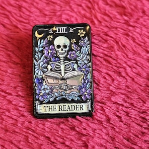 Könnte beinhalten: Rechteckiger Ansteck-Pin mit einem Skelett, das ein Buch liest, umgeben von lila Blumen und Blättern. Der Pin hat einen schwarzen Hintergrund mit dem Text "THE READER" unten. Der Pin befindet sich vor einem rosa, strukturierten Hintergrund.