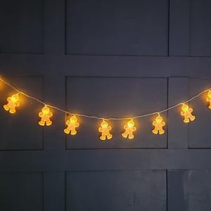 Puede incluir: Una cadena de luces con hombrecitos de jengibre brillantes. Las luces están ensartadas en un cable blanco fino y se alimentan con un pequeño adaptador blanco. Fondo azul oscuro.