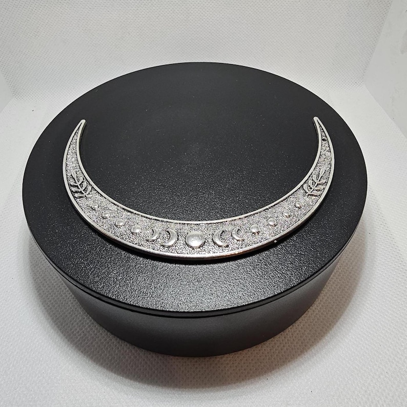 Puede incluir: Una caja redonda negra con un dise&ntilde;o de luna creciente plateada. La luna est&aacute; decorada con peque&ntilde;os c&iacute;rculos y lunas crecientes.