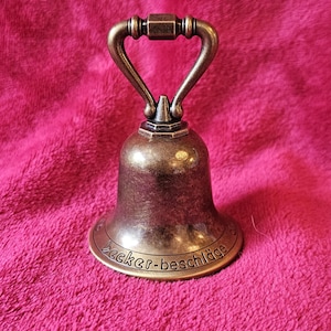 K&ouml;nnte beinhalten: Bronzefarbene Glocke mit gebogenem Griff. Die Glocke hat einen ausgestellten Sockel mit dem Text "Becker-beschl&auml;ge" am unteren Rand. Die Glocke befindet sich vor einem rosa, strukturierten Hintergrund.