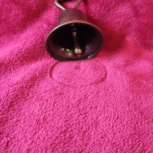 K&ouml;nnte beinhalten: Eine kleine Glocke im antiken Stil mit dunklem Metallfinish. Die Glocke hat einen Schlaufengriff und einen Kl&ouml;ppel im Inneren. Die Glocke befindet sich vor einem leuchtend rosa Hintergrund.