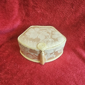Könnte beinhalten: Eine fünfeckige Aufbewahrungsbox, bezogen mit cremefarbenem Stoff mit einem floralen Damastmuster in Ton-in-Ton-Optik. Der Deckel ist mit einer dekorativen gedrehten Kordel umrandet und besitzt einen zentralen Kordelknoten mit Quaste.