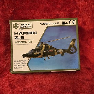 Könnte beinhalten: Modellbausatz für einen Harbin Z-9 Hubschrauber. Die grüne und schwarze Box zeigt "HARBIN Z-9 MODEL KIT" und "BUILD & PLAY". Der Hubschrauber ist in Tarnfarben dargestellt. Maßstab 1:65.
