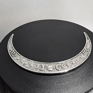 Puede incluir: Un colgante de plata en forma de media luna con un dise&ntilde;o detallado de lunas m&aacute;s peque&ntilde;as y hojas. El colgante est&aacute; sobre un fondo negro.