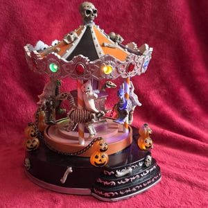 May include: A Halloween-themed carousel decoration featuring a black and orange roof topped with a skull. The ride includes figures of a ghost and skeletons on poles, illuminated by colorful LED lights, and is decorated with small orange jack-o'-lanterns.