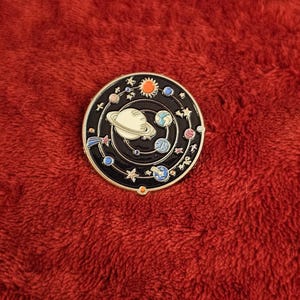May include: A round, black enamel pin featuring a white planet with rings, surrounded by planets, stars, and a sun. The pin is set against a red, textured background.