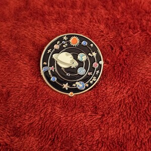 May include: A round, black enamel pin featuring a solar system design. The pin has a white planet with rings, planets, stars, and a sun. The pin is set against a red, textured background.