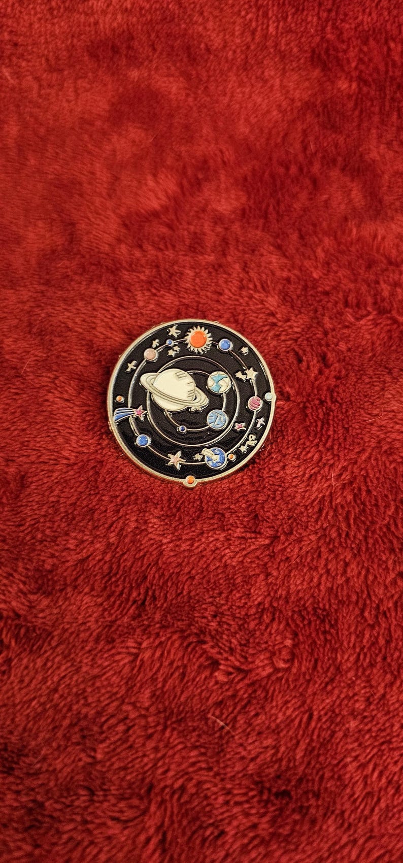 May include: A round, black enamel pin featuring a solar system design. The pin has a white planet with rings, planets, stars, and a sun. The pin is set against a red, textured background.