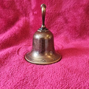K&ouml;nnte beinhalten: Eine bronzefarbene Glocke mit gebogenem Griff und ausgestelltem Sockel. Die Glocke hat ein dekoratives Design um den Sockel und steht auf einer rosa, strukturierten Oberfl&auml;che. Die Glocke wird wahrscheinlich zum Signalisieren oder Dekorieren verwendet.