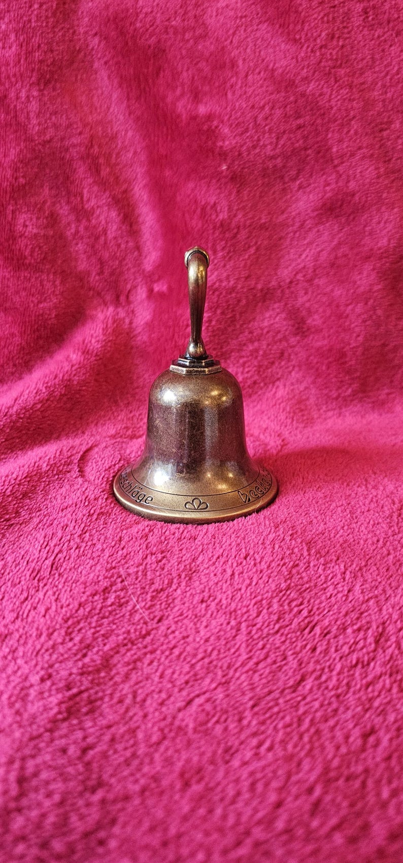K&ouml;nnte beinhalten: Eine bronzefarbene Glocke mit gebogenem Griff und ausgestelltem Sockel. Die Glocke hat ein dekoratives Design um den Sockel und steht auf einer rosa, strukturierten Oberfl&auml;che. Die Glocke wird wahrscheinlich zum Signalisieren oder Dekorieren verwendet.