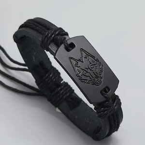 Op de afbeelding: Zwart leren armband met een zwarte metalen wolfskop bedel. De armband is gemaakt van gevlochten leer en heeft een geweven ontwerp.