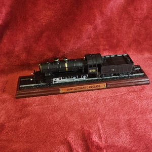 以下が含まれることがあります： 木製の台座に載った、黒いSaint-Gothard C 4/5 Classの模型列車。金色のアクセントと番号2701が特徴です。赤い表面に展示されています。「SAINT-GOTHARD C 4/5 CLASS」と書かれたプレートが付いています。