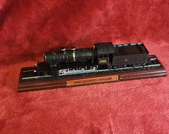 May include: A black model train, the Saint-Gothard C 4/5 Class, sits on a wooden base with a small metal plaque. The train has gold accents and the number 2701 on the side. The model is displayed on a red surface.