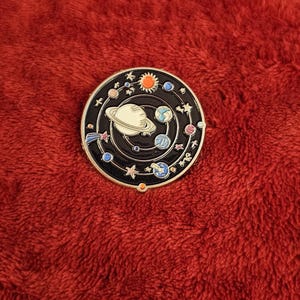 May include: A round, black enamel pin featuring a colorful solar system design. The pin depicts planets, stars, and the sun, with a white Saturn at the center. The pin is set against a red, textured background.