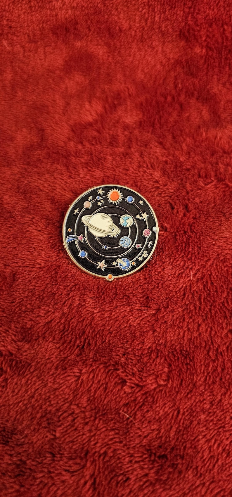 May include: A round, black enamel pin featuring a colorful solar system design. The pin depicts planets, stars, and the sun, with a white Saturn at the center. The pin is set against a red, textured background.