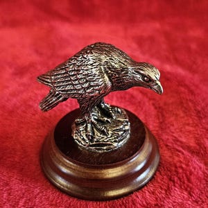 Könnte beinhalten: Detaillierte silberfarbene Adlerfigur auf einem dunklen Holzsockel. Der Adler ist mit aufwendigen Federdetails und einem fokussierten Blick gestaltet. Der runde Sockel hat eine polierte Oberfläche, vor rotem Hintergrund.