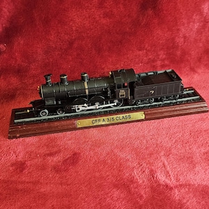 May include: A detailed model of a black steam train, mounted on a wooden base with a brass nameplate. The train features intricate detailing, including wheels, smokestacks, and a tender car. The nameplate reads "CFF A 3/5 CLASS".