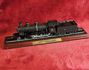 May include: A detailed model of a black steam train, mounted on a wooden base with a brass nameplate. The train features intricate detailing, including wheels, smokestacks, and a tender car. The nameplate reads "CFF A 3/5 CLASS".