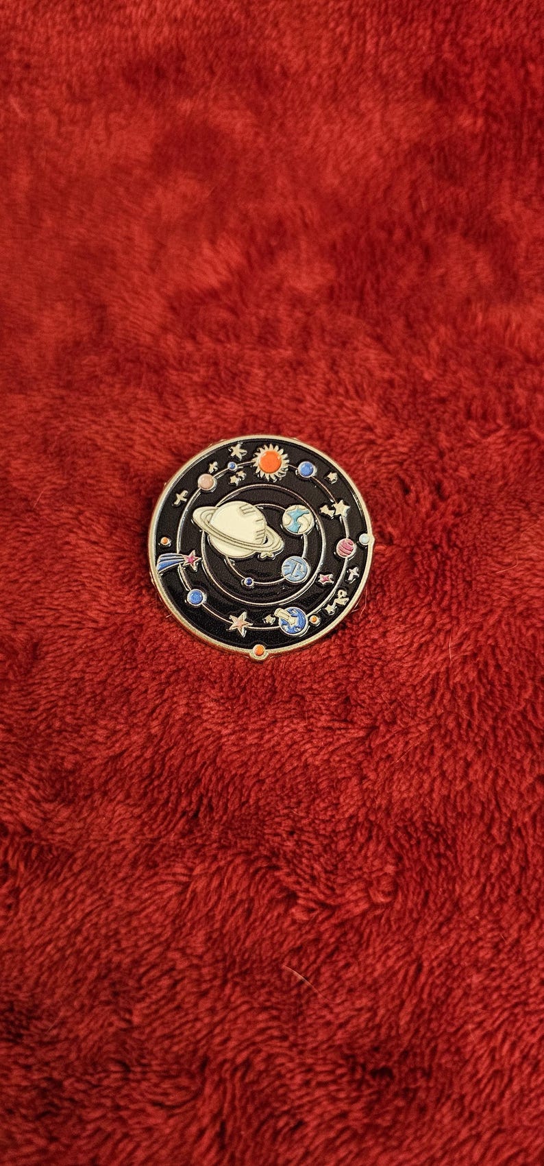 May include: A round, black enamel pin featuring a colorful solar system design. The pin includes planets, stars, and a sun, with a central planet ringed in white. The pin is set against a red, textured background.