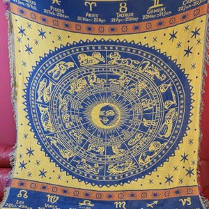 Könnte beinhalten: Eine gelb-blaue Decke mit einem Tierkreis-Rad-Design. Die Decke hat ein zentrales kreisförmiges Design mit Tierkreiszeichen und Text, umgeben von Sternen. Die Ränder haben eine Fransenborte.