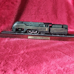 May include: A model of an SNCB 12 Class locomotive, in dark green and black, on a wooden base with tracks. The model train has detailed wheels and is labelled "SNCB 12 CLASS".