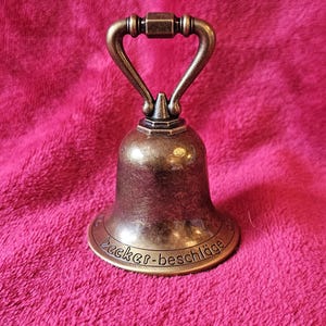K&ouml;nnte beinhalten: Eine bronzefarbene Glocke mit gebogenem Griff und ausgestelltem Sockel. Die Glocke tr&auml;gt den Text "Becker-beschl&auml;ge" am Sockel. Die Glocke befindet sich vor einem rosa, strukturierten Hintergrund.