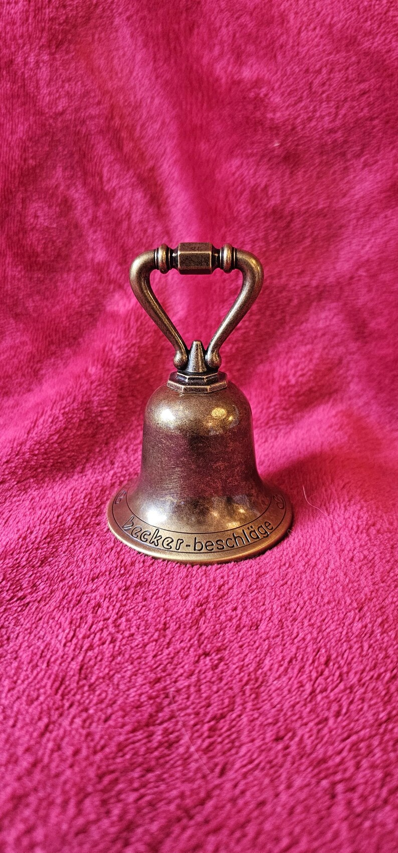K&ouml;nnte beinhalten: Eine bronzefarbene Glocke mit gebogenem Griff und ausgestelltem Sockel. Die Glocke tr&auml;gt den Text "Becker-beschl&auml;ge" am Sockel. Die Glocke befindet sich vor einem rosa, strukturierten Hintergrund.