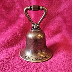 K&ouml;nnte beinhalten: Eine bronzefarbene Glocke mit gebogenem Griff und dem Text "Becker-beschl&auml;ge" am Sockel. Die Glocke befindet sich vor einem rosa, strukturierten Hintergrund.