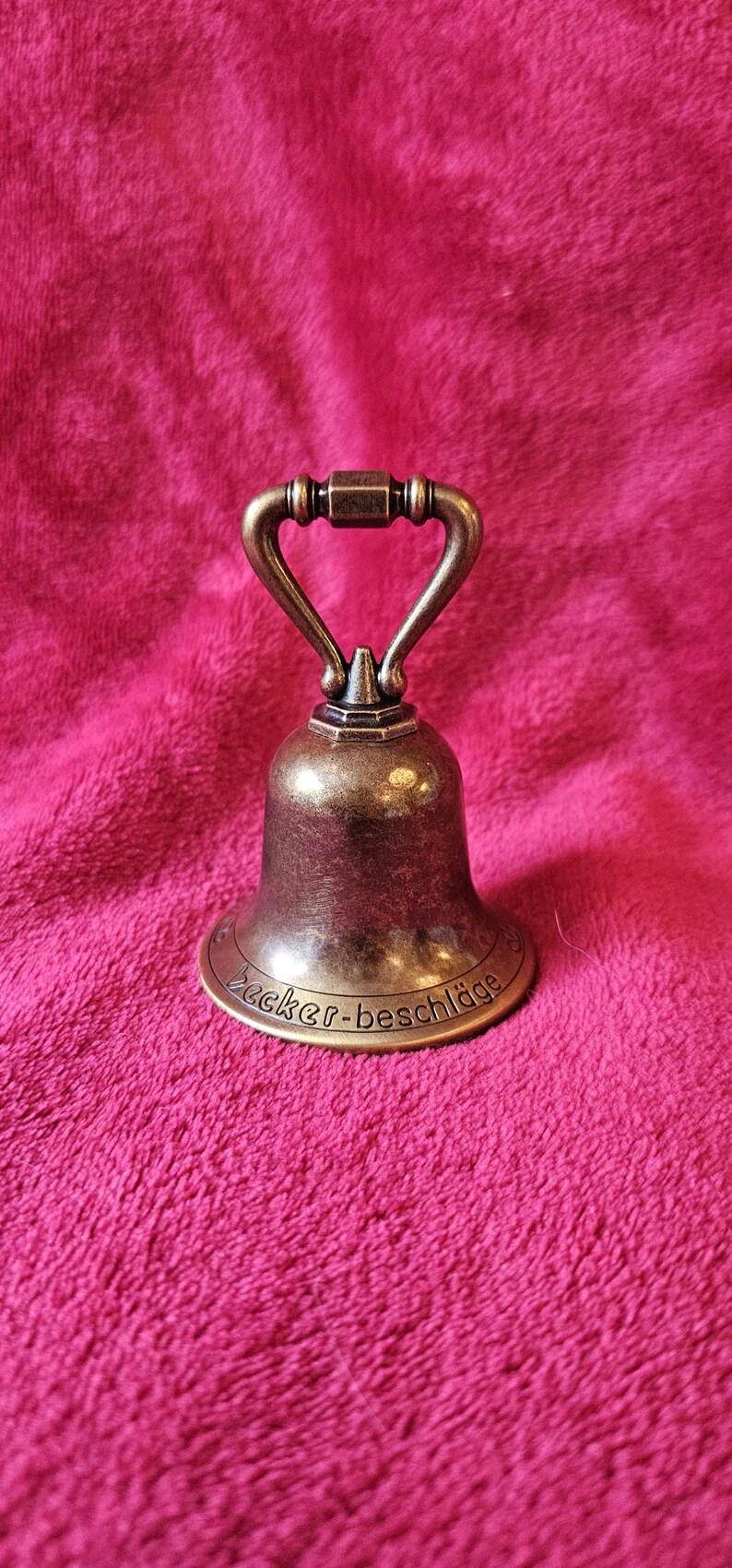 K&ouml;nnte beinhalten: Eine bronzefarbene Glocke mit gebogenem Griff und dem Text "Becker-beschl&auml;ge" am Sockel. Die Glocke befindet sich vor einem rosa, strukturierten Hintergrund.