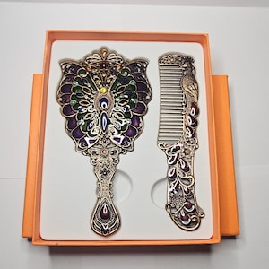 Peut inclure: Un ensemble de deux accessoires pour cheveux dorés dans une boîte. Le premier accessoire est un miroir de poche avec un motif de papillon et des accents violets, verts et dorés. Le deuxième accessoire est un peigne avec un motif de paon et des accents rouges, dorés et violets.