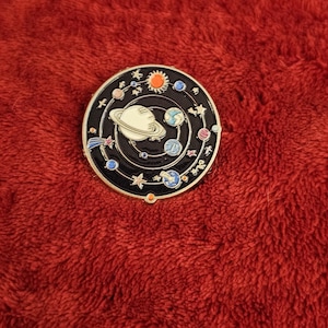 May include: A round, black enamel pin featuring a colorful solar system design. The pin includes planets, stars, and a sun, all set against a black background. The pin is on a red, textured surface.