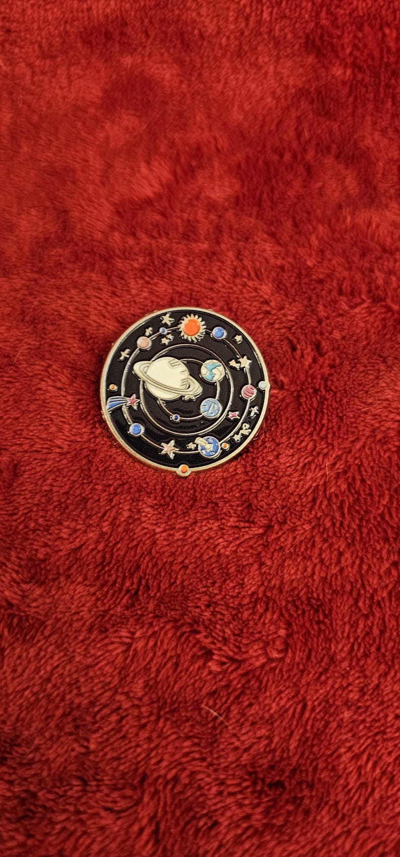 May include: A round, black enamel pin featuring a colorful solar system design. The pin includes planets, stars, and a sun, all set against a black background. The pin is on a red, textured surface.