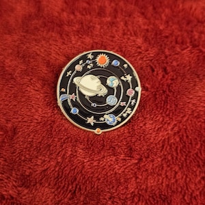 May include: A circular, black enamel pin depicting a white planet with rings, encircled by planets, stars, and a sun. The pin is set against a red, textured backdrop.