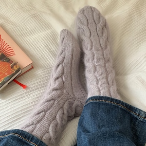 Può includere: Un paio di calzini morbidi in maglia a treccia di colore lilla chiaro con una texture soffice. I calzini sono indossati ai piedi di una persona e riposano su un copriletto bianco.