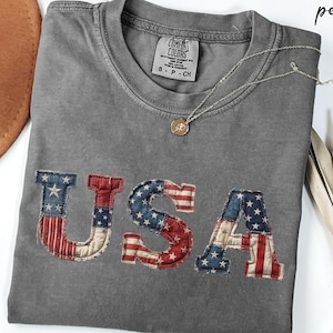 Puede incluir: Una camiseta gris con la palabra "USA" aplicada en la parte delantera con un diseño de bandera patriótica. La camiseta tiene cuello redondo y mangas cortas. Un collar dorado con un colgante está drapeado sobre la camiseta.