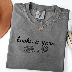 May include: A gray t-shirt with the words "books & yarn" in black script, along with a graphic of stacked books and a ball of yarn. A gold necklace with a pendant rests on the shirt. The shirt is folded on a white surface.