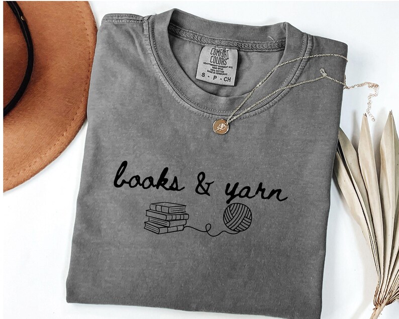 May include: A gray t-shirt with the words "books & yarn" in black script, along with a graphic of stacked books and a ball of yarn. A gold necklace with a pendant rests on the shirt. The shirt is folded on a white surface.