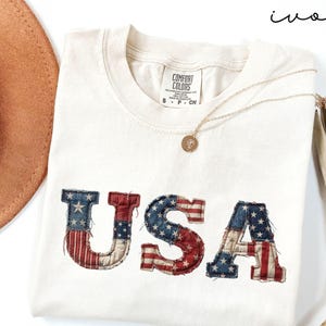 May include: Ivory-colored t-shirt with the word "USA" in a patriotic design. The letters are in red, white, and blue, resembling the American flag. A gold necklace and a brown hat are also visible.