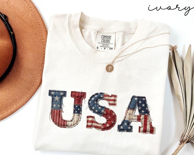 May include: Ivory-colored t-shirt with the word "USA" in a patriotic design. The letters are in red, white, and blue, resembling the American flag. A gold necklace and a brown hat are also visible.