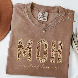 May include: A brown t-shirt with the words "MOH" in gold glitter letters with a leopard print pattern. The words "maid of honor" are written in cursive below the letters.