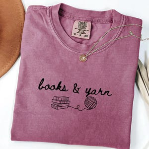 May include: A mauve Comfort Colors t-shirt with the words "books & yarn" in black script, along with a graphic of books and yarn. A gold necklace with a pendant rests on the shirt. A brown hat and dried plant are in the background.