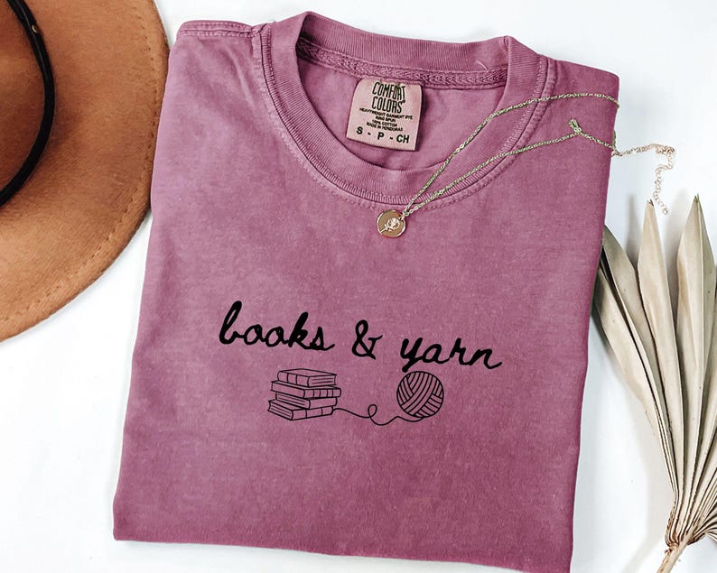 May include: A mauve Comfort Colors t-shirt with the words "books & yarn" in black script, along with a graphic of books and yarn. A gold necklace with a pendant rests on the shirt. A brown hat and dried plant are in the background.