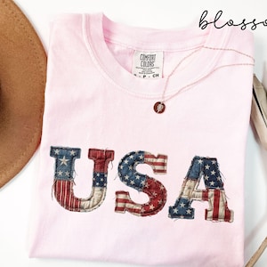 May include: A light pink t-shirt with the word "USA" in a distressed American flag design. The shirt has a crew neck and a small tag on the collar. A gold necklace with a pendant is draped across the shirt.