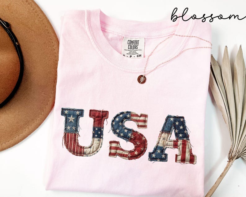 May include: A light pink t-shirt with the word "USA" in a distressed American flag design. The shirt has a crew neck and a small tag on the collar. A gold necklace with a pendant is draped across the shirt.