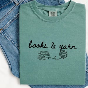 May include: A seafoam green t-shirt with the words "books & yarn" in black script, along with a graphic of stacked books and a ball of yarn. The shirt is displayed on a pair of blue jeans.