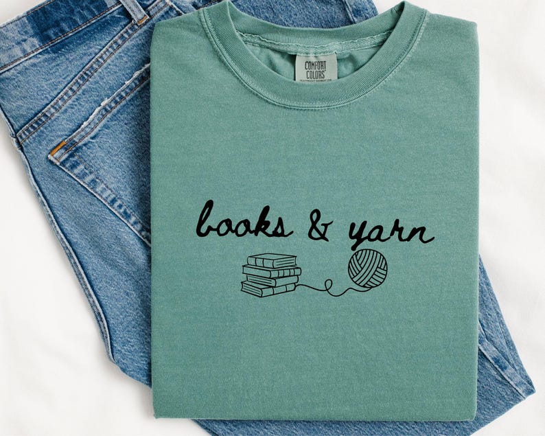 May include: A seafoam green t-shirt with the words "books & yarn" in black script, along with a graphic of stacked books and a ball of yarn. The shirt is displayed on a pair of blue jeans.