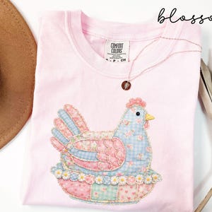May include: A light pink t-shirt featuring a patchwork chicken design in shades of blue, pink, and green. The chicken is sitting on a nest with a floral trim. A gold necklace with a pendant is draped on the shirt.