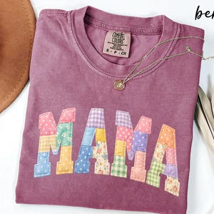 May include: A berry-colored Comfort Colors t-shirt with the word "MAMA" appliqued in colorful patchwork fabric. A gold necklace with a pendant rests on the shirt. The shirt is folded on a white surface.